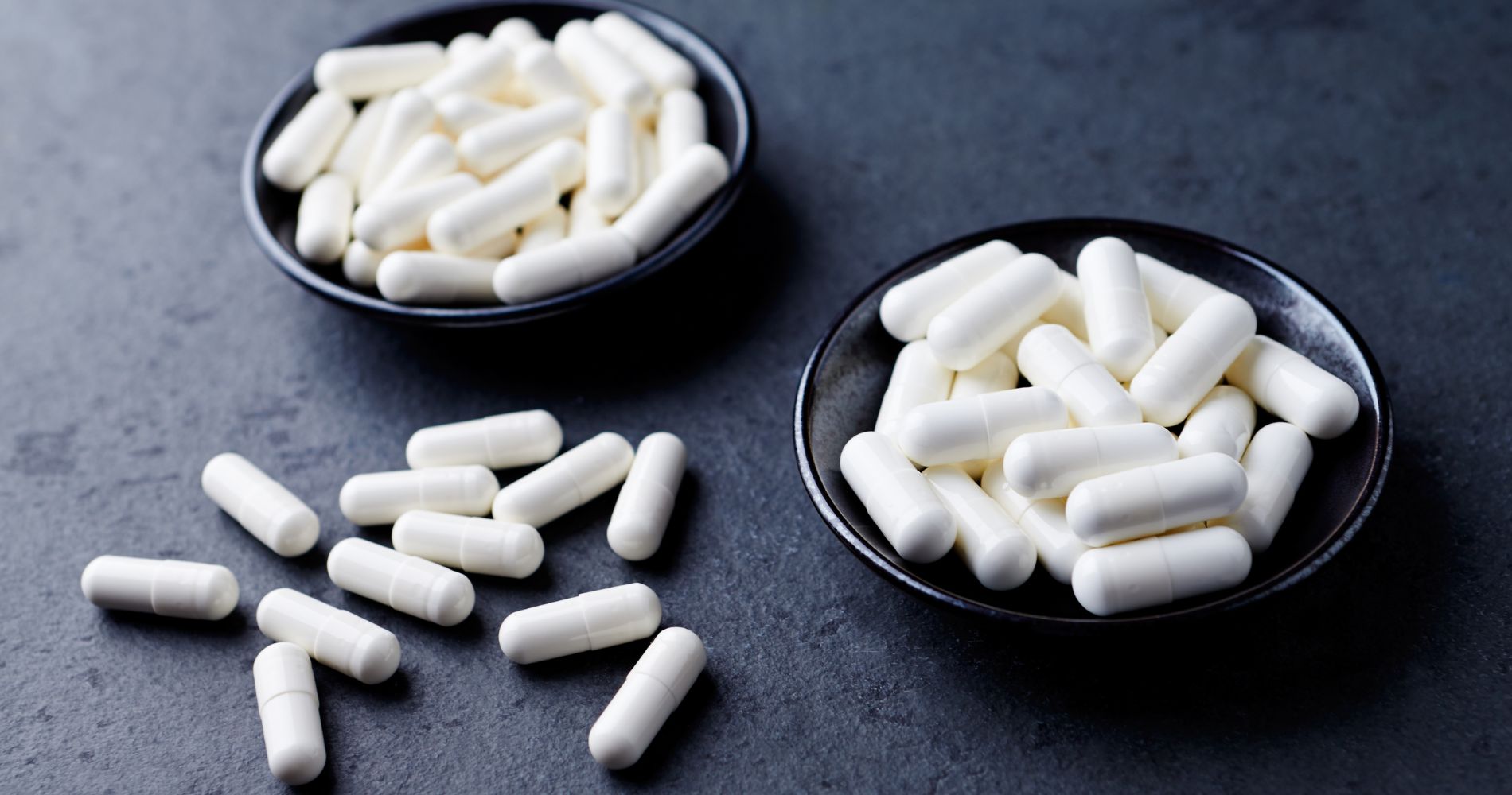 Białe kapsułki z argininą w miseczkach na ciemnym tle – suplementacja L-argininą w formie doustnej.